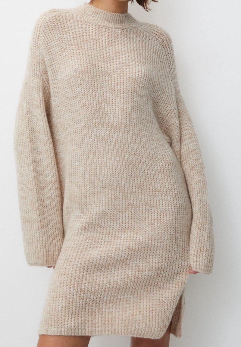 Femme portant une robe-pull en maille beige à manches longues avec une fente sur le côté, debout devant un fond blanc uni.