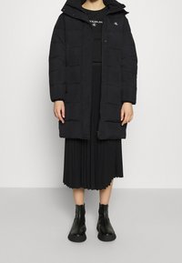 Černá prošívaná bunda s vysokým límcem. Oblékaná přes černou plisovanou sukni a černé kotníkové boty.