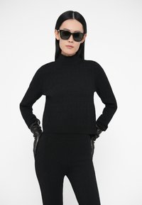 Mustast kudumist kõrge kaelusega kampsun tekstuurse mustriga ja soonikkoes äärega, kombineeritud mustade kinnaste ja päikeseprillidega, valgel taustal.