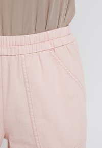 Světle růžové kalhoty s elastickým pasem, bočními kapsami a viditelným šitím. Materiál má hladkou strukturu s mírným leskem.