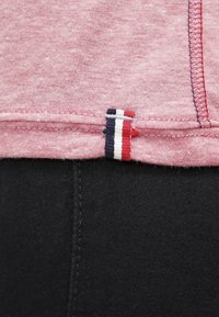 Röd och vit randig tygetikett fäst vid en rosa texturerad skjorta, med svart tyg synligt nedanför. Sömmar är synliga längs fållen.