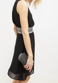 Mujer que lleva un vestido negro sin mangas con cinturón de cintura estampado, sosteniendo un pequeño bolso negro texturizado en su mano derecha.