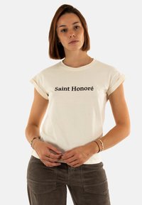 T-shirt in cotone crema con maniche arrotolate, caratterizzato da testo nero "Saint Honoré" posizionato centralmente. Tessuto morbido, vestibilità rilassata.