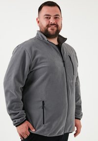 Sivý fleeceový kabát so zipsom vpredu, vysokým golierom a dvoma bočnými vreckami so zipsom. Mäkka textúra a uvoľnený strih.