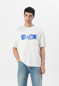 Camiseta blanca de manga corta con un audaz gráfico azul de "HugO". Hecha de algodón suave, con un corte relajado y escote redondo.