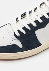 Białe sneakersy z ciemnoniebieskimi zamszowymi akcentami, perforowanym noskiem, beżową podeszwą i beżowymi sznurowadłami, charakteryzujące się smukłym, minimalistycznym designem.