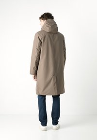 Lang brun frakk med hette, jevnt stoff, lengre silhuett, sidesømmer og bakventil; brukt med mørk denim jeans og hvite sko.