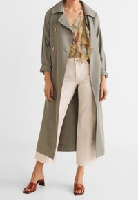 Oliwkowy trencz z podwójnymi guzikami, nałożony na kwiatową bluzkę, w połączeniu z kremowymi szerokimi spodniami i brązowymi sandałami na obcasie.