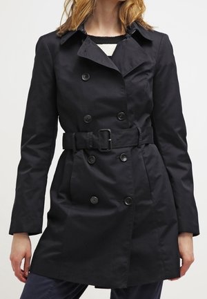 Mujer con gabardina negra de doble botonadura y cinturón, de pie con las manos relajadas a los lados sobre un fondo liso.