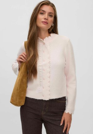 Jeune femme aux longs cheveux bruns portant un cardigan blanc à bords festonnés, un pantalon foncé et tenant un sac à bandoulière en daim beige.