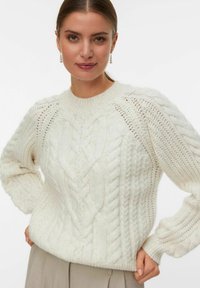 Vrouw in een wit kabelgebreid vest met lange pofmouwen, gekleed in lichtbeige pantalons, staande met handen op de heupen.
