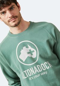 Sudadera de algodón verde con un gráfico central en blanco y el texto "ALTONADOCK es tu camino." Textura suave y diseño de cuello redondo.