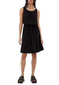 Frau in einem ärmellosen schwarzen knielangen Kleid mit einem gebundenen Taillengürtel und schwarzen Plateausandalen, steht vor einem weißen Hintergrund.