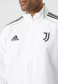 Hvid træningsjakke med sorte og grønne detaljer. Har Adidas-logo og Juventus-emblem. Høj krave, lynlås foran og stribede skuldre.