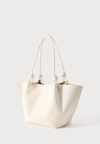 Krémově zbarvená kožená kabelka s dvojitými provazovými rukojeťmi uvázanými u zlatých kovových průvleků, strukturované měkké tělo, na bílém pozadí.