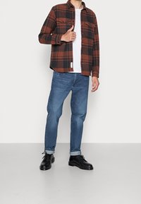 Červeno-černá kostkovaná flanelová košile přes bílý tričko, spárovaná se světle modrými džíny a černými �šněrovacími botami. Košile má dvě náprsní kapsy.