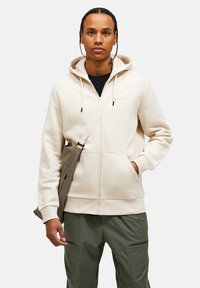 Creme zip-up hoodie med en frontlomme, snorhue og broderet logo. Båret med mørke bukser og en beige messenger bag.