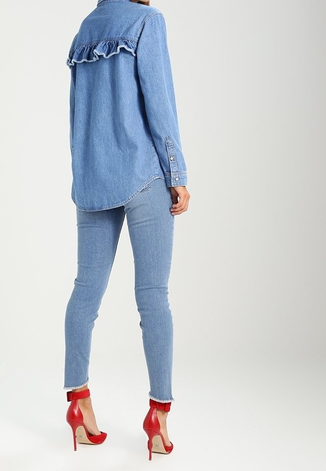 Femme portant une chemise en denim bleu clair avec un détail volanté au dos, un jean skinny assorti et des escarpins rouges à brides à la cheville, debout.