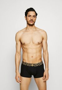 Svarte boxerbriefs med en gyllen mønstret linning, som viser en jevn tekstur og tettsittende design, modellert av en barbrystet mannlig modell.