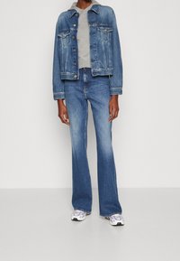 Denim takki napin kiinnityksellä, harmaa pyöreäkauluksinen paita, levenevät siniset farkut ja valkoiset tennarit violetilla yksityiskohdalla. Rentoa istuvuutta, arkinen texture.