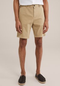Pantaloni scurți din bumbac bej, cu croi drept, închidere cu buton în față și bucle pentru curea. Purtați cu pantofi slip-on țesuti negri. Fundal simplu.