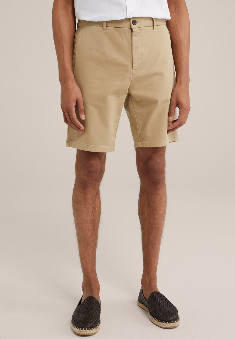 Pantaloni scurți din bumbac bej, cu croi drept, închidere cu buton în față și bucle pentru curea. Purtați cu pantofi slip-on țesuti negri. Fundal simplu.
