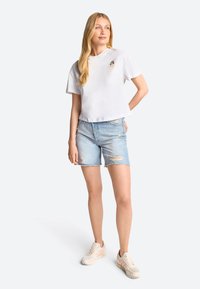 White cropped t-shirt with a small embroidered detail, paired with light blue distressed denim shorts, and beige athletic shoes.
