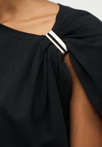 Κοντινό πλάνο ενός ατόμου που φοράει μαύρο τοπ με μανίκια και διακοσμητικό κούμπωμα διπλής μπάρας σε χρυσό τόνο στον ώμο.