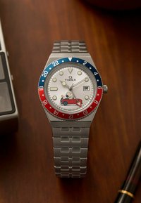 Montre Timex Q avec un bracelet en acier inoxydable argenté, cadran blanc rond, lunette rouge et bleue, et design de personnage de cartoon dans une voiture.