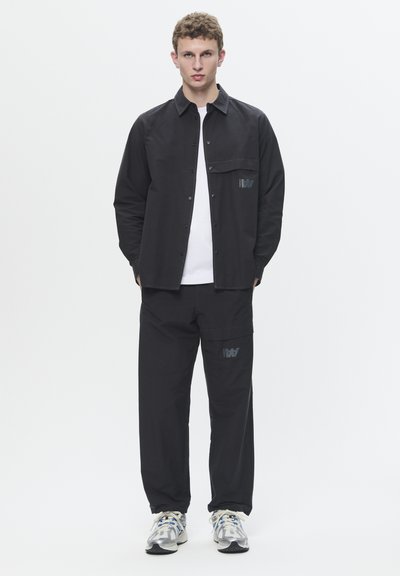Schwarzes Hemd mit Knopfleiste und Brusttasche, kombiniert mit passenden schwarzen Cargo-Hosen mit einem Logo. Helle Sportschuhe mit Akzenten.