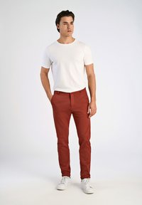 Pantaloni rossi testurizzati con una vestibilità slim, abbinati a una t-shirt bianca a maniche corte e sneakers bianche. Il modello sta in piedi con una mano in tasca.