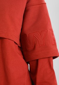 Camisa corta de algodón roja con un diseño en capas y un logo bordado "NY" en un color a juego en la manga. Textura suave.