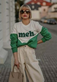 Zelený a bílý svetr s barevnými bloky a textem "meh.", doplněný b éžovými kalhotami s vysokým pasem a malou béžovou kabelkou.