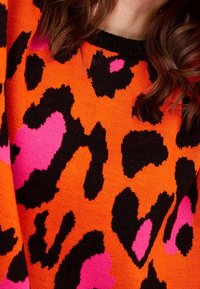 Pull en maille avec un fond orange orné de taches de léopard noires audacieuses et d'accents roses vifs. Tissu texturé avec un col rond.