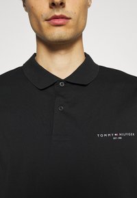 Gros plan sur une personne portant un polo noir Tommy Hilfiger avec deux boutons et le logo sur la poitrine gauche.