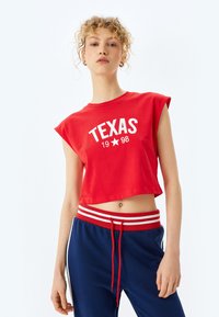 Rød cropped t-shirt med "TEXAS 1998" trykt i hvidt, med cap ærmer. Sammen med navy sweatpants med en rød og hvid stribet talje.