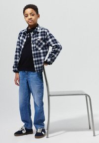 Blauwe en witte geruite flanellen shirt met een zwart T-shirt, lichtblauwe jeans en donkere sneakers, staande naast een zilveren stoel.