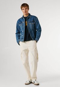 Kurtka dżinsowa w ciemnym niebieskim kolorze, zestawiona z czarnym t-shirtem z żółtym nadrukiem, białymi spodniami typu cargo oraz białymi trampkami z czarnymi akcentami.