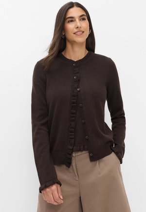 Vrouw met lang bruin haar draagt een donkerbruine gebuttonde cardigan met ruchezoom en beige wijde broek, staand tegen een witte achtergrond.