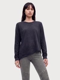 Giovane donna con capelli lisci e scuri che indossa un top nero asimmetrico a maniche lunghe e pantaloni grigi con trama, su uno sfondo semplice.