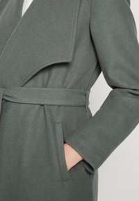 Lång, grå ullkappa med sjalkrage, belagd midja och sidofickor. Texturen ser slät och varm ut, med en minimalistisk design.