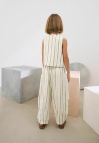 Vit randig tvådelad outfit med ärmlös topp och löst sittande byxor. Slät textur, bärs med bruna skor. Enkla bakgrund med geometriska former.