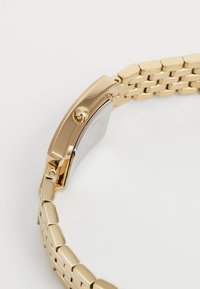 Montre-bracelet dorée avec un boîtier rectangulaire, un bracelet en maillons métalliques et une couronne ronde. Surface lisse avec finition polie.