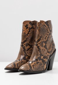 Botas de tobillo con estampado de serpiente en tonos marrón y beige, con puntera afilada, cremallera lateral y tacón grueso, que presentan una textura brillante.