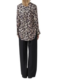 Femme portant une blouse beige et noire à imprimé léopard à manches longues, non rentrée, sur un pantalon noir à jambes larges, dos à l'appareil photo.