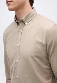 Άνδρας με μπεζ πουκάμισο με κουμπιά και κολάρο με κουμπιά, εμφανίζεται από τον λαιμό μέχρι το μέσο του κορμού σε απλό φόντο.