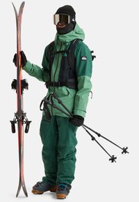 Groene en zwarte skikleding met een waterdichte jas en broek, houdend een paar rode ski's en zwarte skistokken met sneeuwmanden. Grote spiegeltjesgoggles.