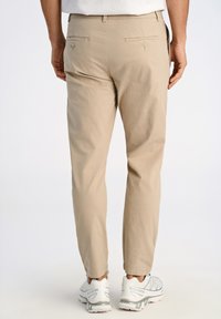 Pantalon en coton beige avec une coupe ajustée, comportant deux poches arrière avec fermetures à bouton. Porté avec des baskets grises et blanches.