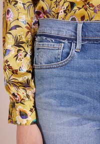 Blusa di seta gialla floreale con maniche a polsino abbinata a jeans di denim azzurri chiari con tasca sfrangiata e gradiente da chiaro a scuro.