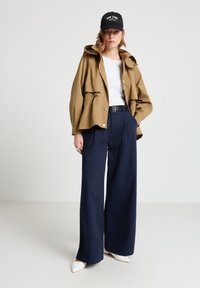 Hellbraune Kapuzenjacke mit Kordelzug über einem weißen Oberteil, kombiniert mit marineblauen, weit geschnittenen Hosen und weißen spitzen Schuhen; schwarze Mütze mit Schriftzug.